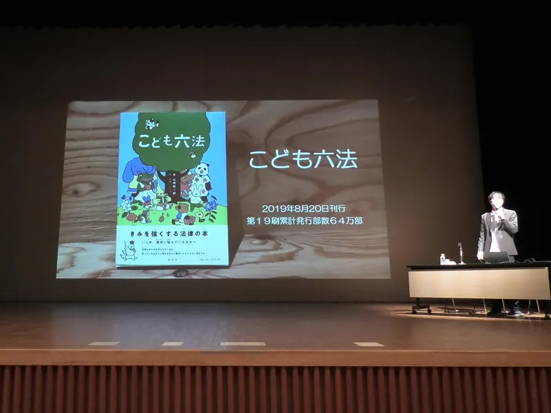 『こども六法』の表紙を大きく映した舞台で講演する山﨑聡一郎