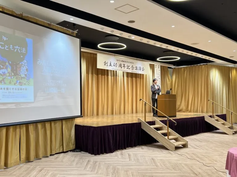 「記念講演会」の横断幕が掲げられたホールの演台で講演する山﨑聡一郎