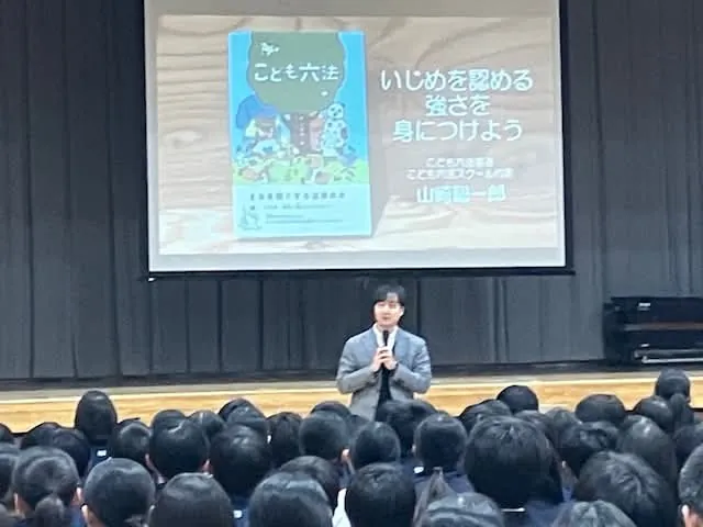 学校の体育館で「いじめを認める強さを身につけよう」のスライドを背に、生徒たちに話す山﨑聡一郎