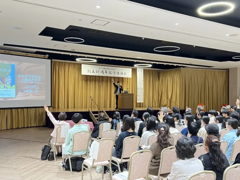 大ホールの講演会で、会場の参加者が手を挙げて問いかけに答えている様子